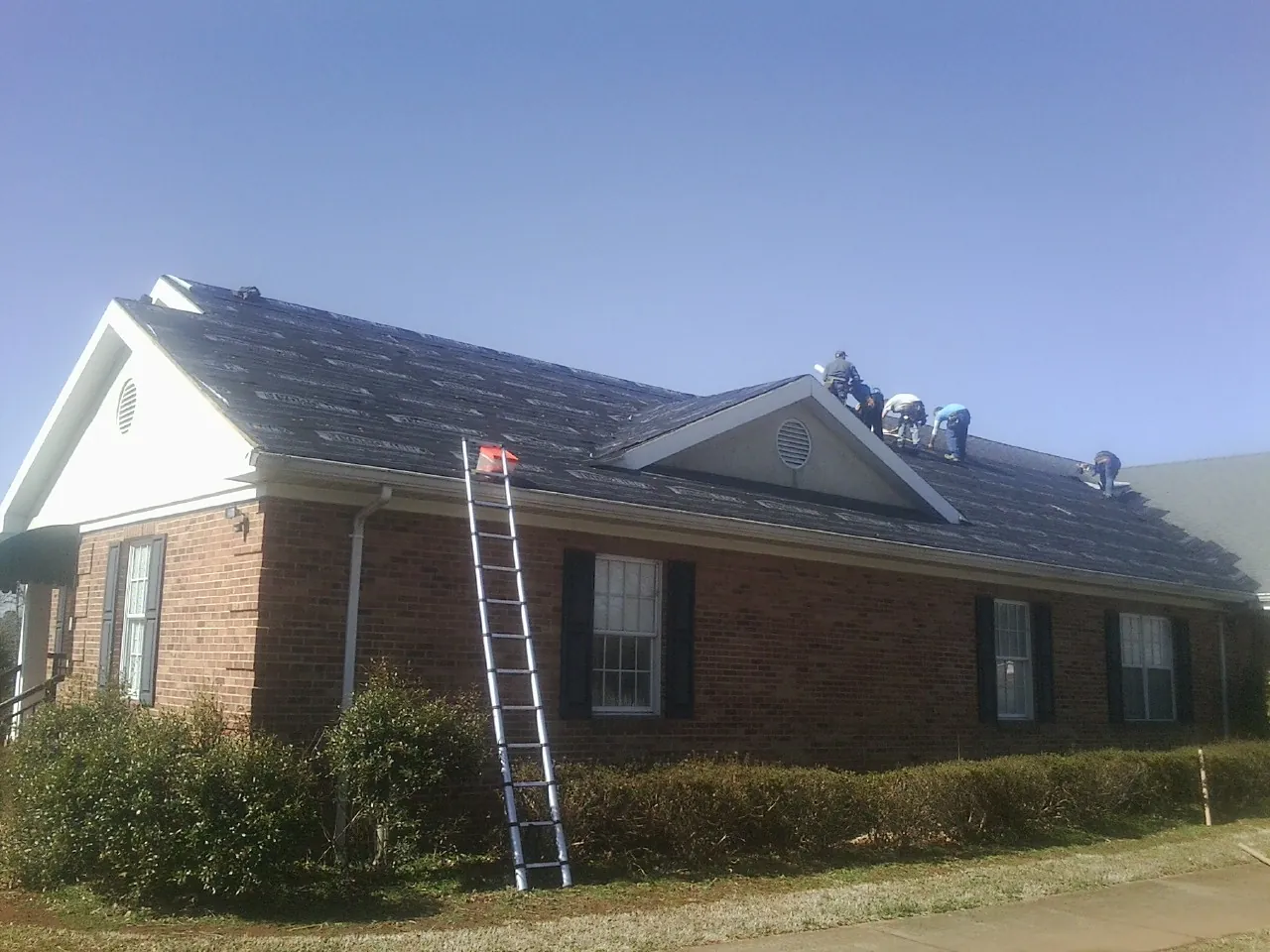 Crew tearing shingles off a brick ranch roof with a ladder set — Metal Roof Repair work by our crew in Topping