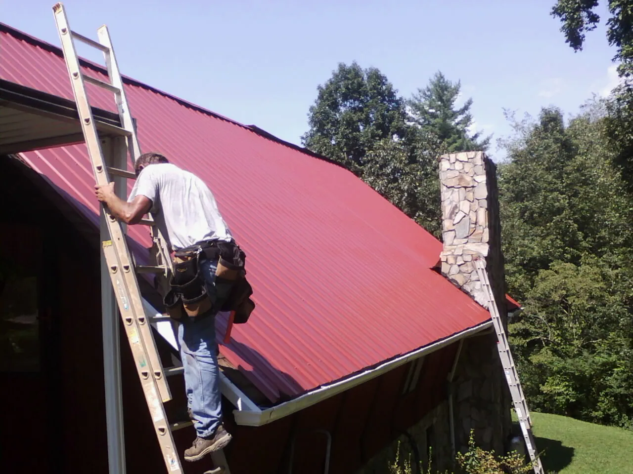 Roofer on a ladder beside a red metal roof and stone chimney — Soffit & Fascia Repair by Roofing Brothers in Topping